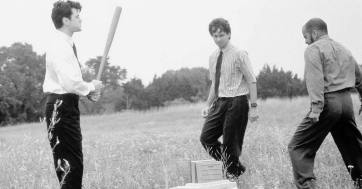 Three men in business attire standing in a field, one holding a baseball bat near a box on the ground, suggesting unconventional or symbolic action outside a traditional workplace.