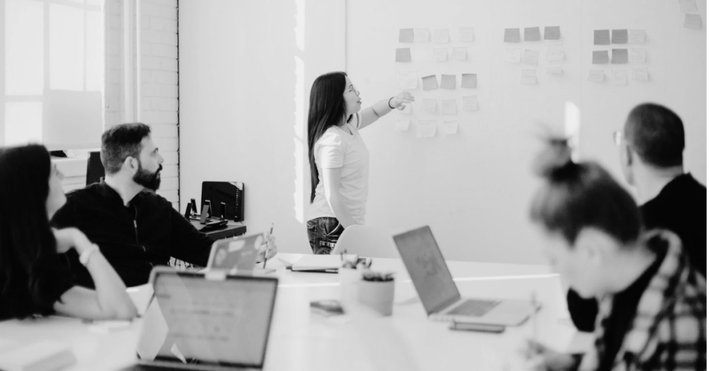 Team meeting in a modern office, with one person presenting ideas on a wall of sticky notes while others watch and work on laptops, illustrating collaboration and planning.