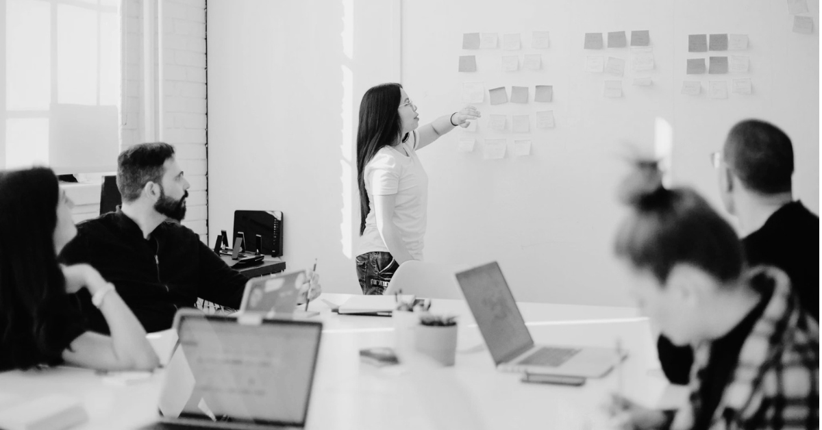 Team meeting in a modern office, with one person presenting ideas on a wall of sticky notes while others watch and work on laptops, illustrating collaboration and planning.