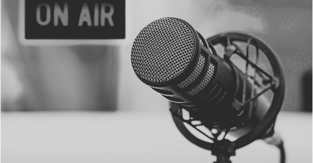 Close-up of a studio microphone in a shock mount with a blurred “On Air” sign in the background, suggesting a live broadcast or recording setting.