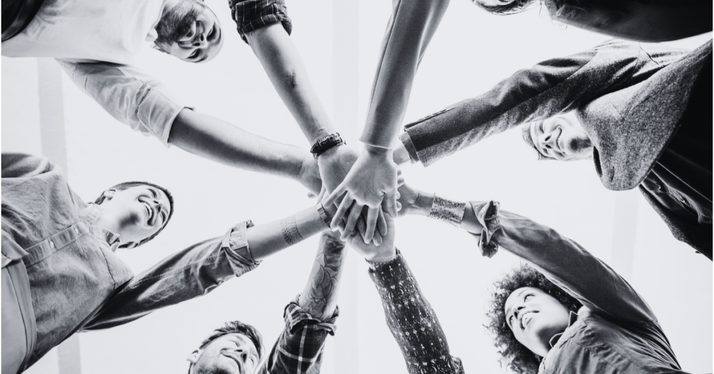 Low-angle view of a diverse group of people standing in a circle with their hands stacked together in the center, symbolizing teamwork and collaboration.