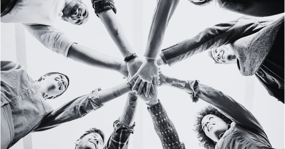 Low-angle view of a diverse group of people standing in a circle with their hands stacked together in the center, symbolizing teamwork and collaboration.