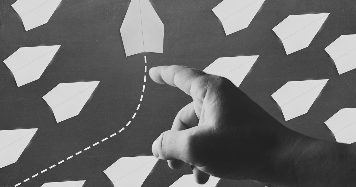 Close-up of a hand pinching a paper airplane among many on a dark surface, with a dashed curved line suggesting launch.