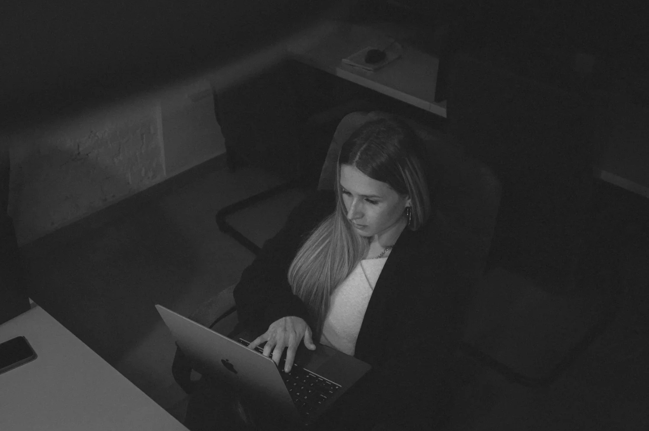 Woman wearing a dark coat typing on a silver laptop at a desk in a dimly lit room.