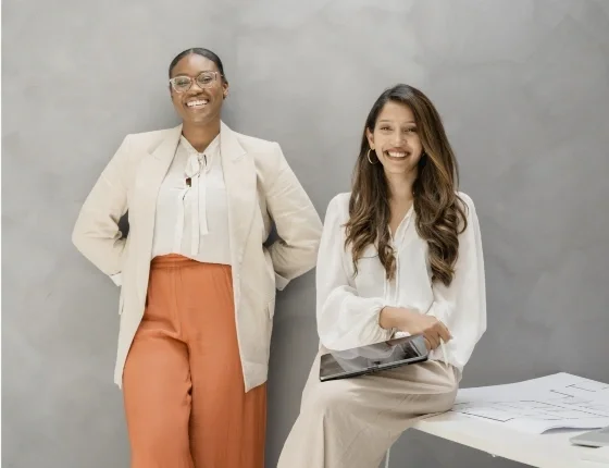 Two professional women posing and smiling in an office setting: one standing in a beige blazer and orange pants, the other seated in a white blouse with a tablet and papers on a desk.